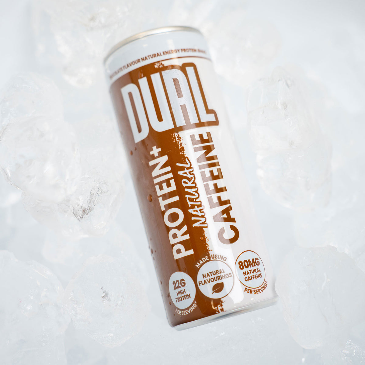 A can of DUAL Protein+ Natural Caffeine drink resting on crushed ice. The can features a white and brown design with bold text, and the background consists of clear ice cubes.
