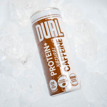A can of DUAL Protein+ Natural Caffeine drink resting on crushed ice. The can features a white and brown design with bold text, and the background consists of clear ice cubes.