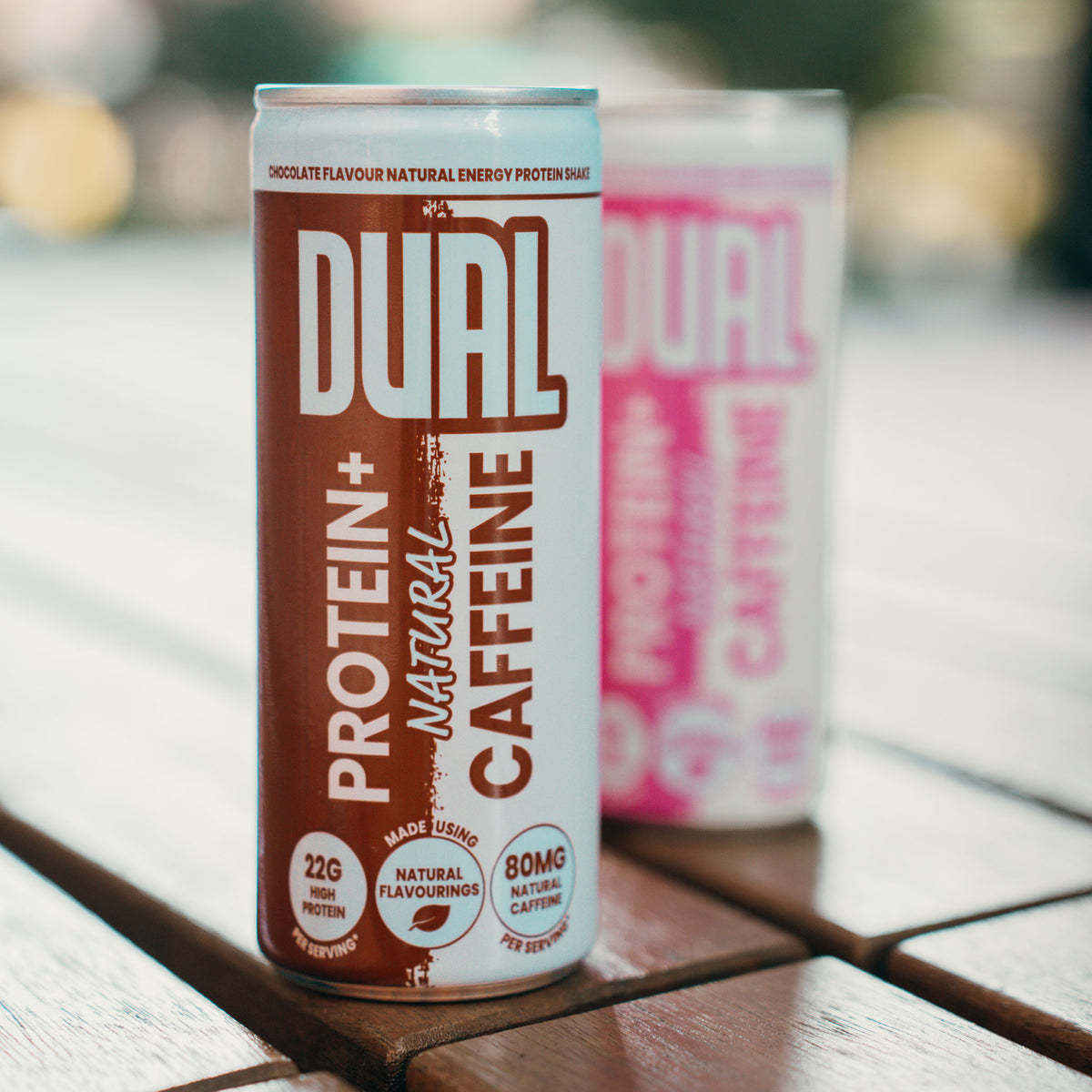 A can of DUAL Protein+ Natural Caffeine drink in chocolate flavour, standing on a wooden table with a second blurred can in the background.