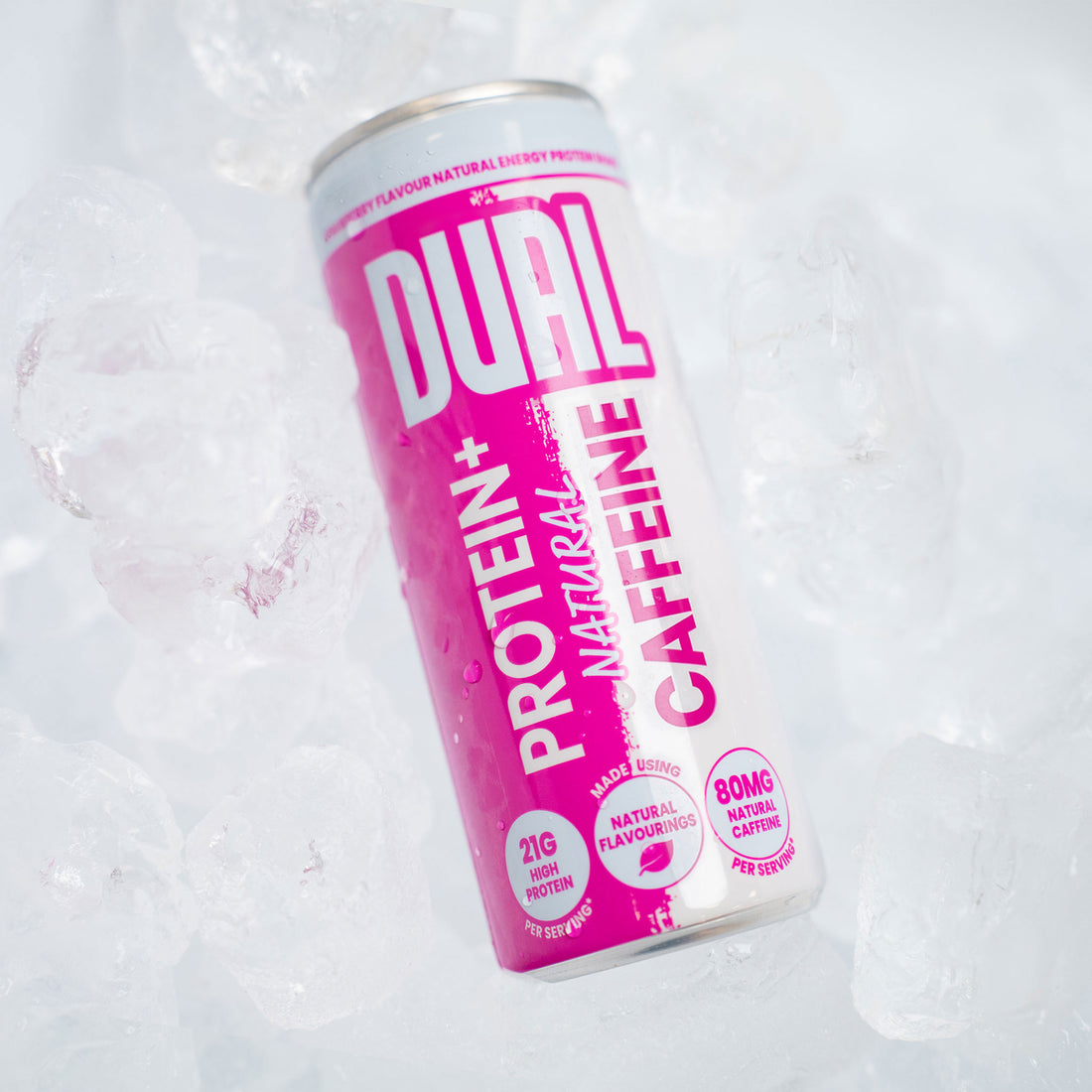 A can of Dual Protein+ Natural Caffeine drink surrounded by ice cubes. The can features a vibrant pink and white label with bold white and pink text.