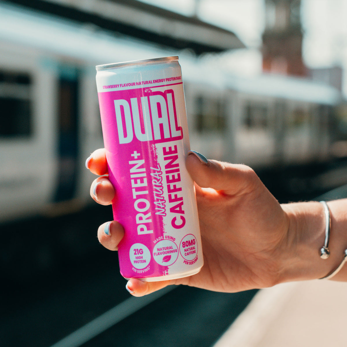 A hand holding a can of DUAL Protein+ Caffeine drink, with a blurred train station platform and train in the background.